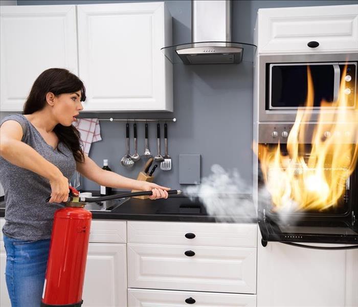 An oven on fire with a women holding a fire extinguisher, pointing it toward the flames.